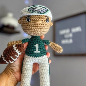以下が含まれることがあります： 緑のジャージに「1」の番号、刺繍された鷲のロゴのついた白い帽子、白いズボンを着用した、かぎ針編みのフットボール選手のドール。ドールは茶色と白のかぎ針編みのフットボールを持っています。