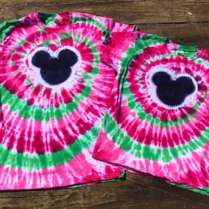 Könnte beinhalten: Zwei Batik-T-Shirts mit einer schwarzen Mickey-Maus-Silhouette auf jedem. Die Shirts sind pink, grün und weiß.