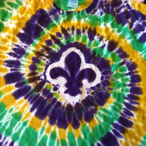 May include: Tie-dye t-shirt in green, gold, and purple, featuring a white and purple fleur-de-lis design at the center. The shirt has a crew neck and short sleeves. The tie-dye pattern radiates from the center.