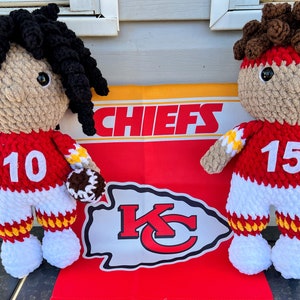 May include: Two crocheted football player dolls wearing red and white jerseys with the numbers 10 and 15. The dolls are holding footballs and are standing in front of a Chiefs flag.