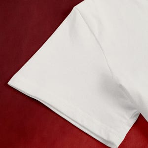 May include: Close-up of a white t-shirt sleeve against a deep red background. The sleeve is made of a smooth, opaque fabric, with visible stitching along the hem and seams. The contrast between the white fabric and the red backdrop creates a striking visual.