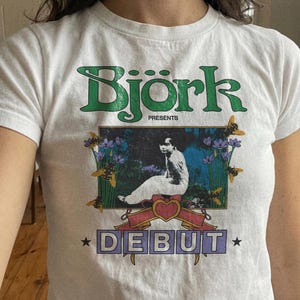 May include: White t-shirt with the word "Bjork" in green, and the word "Debut" in a purple box. The graphic includes a person, bees, and flowers. The shirt has a crew neck and short sleeves.