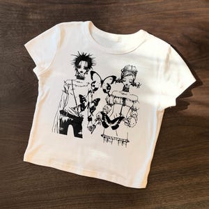 May include: White t-shirt featuring a black and white illustration of two figures with butterfly accents. The graphic design has a manga or anime style. The shirt is made of a soft, comfortable material.