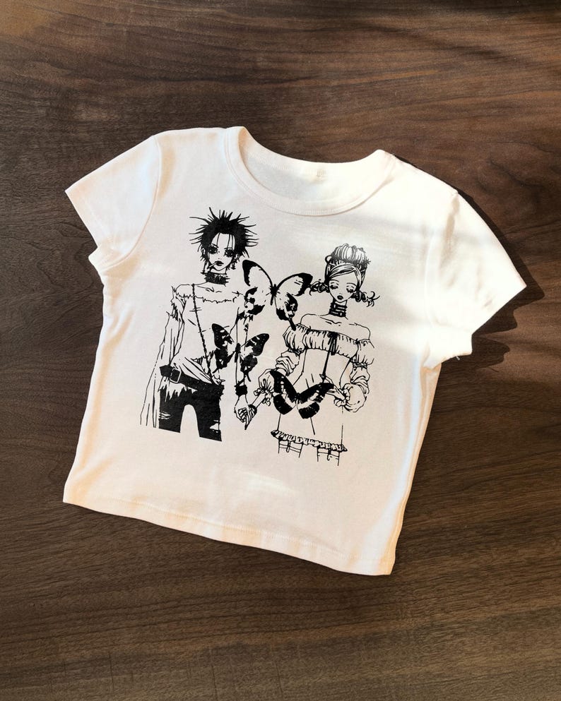 May include: White t-shirt featuring a black and white illustration of two figures with butterfly accents. The graphic design has a manga or anime style. The shirt is made of a soft, comfortable material.