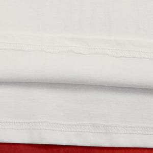 May include: Close-up of white fabric with visible stitching along the seams. The fabric is folded over, revealing a second layer. The background is a solid red color. The image highlights the texture and construction of the textile.