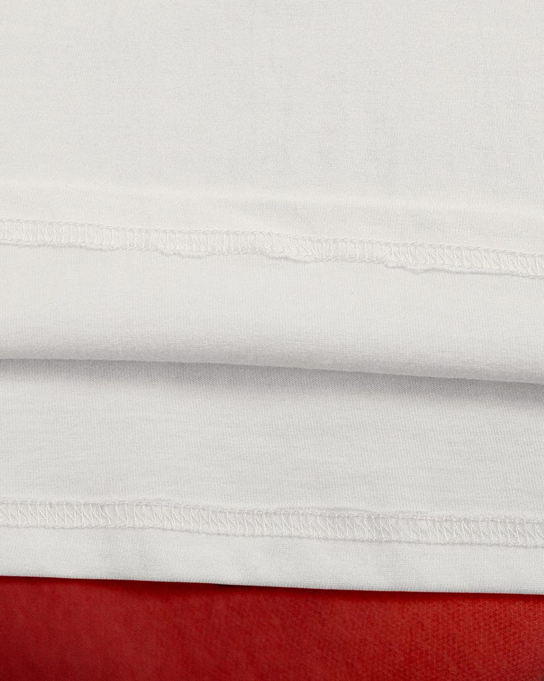 May include: Close-up of white fabric with visible stitching along the seams. The fabric is folded over, revealing a second layer. The background is a solid red color. The image highlights the texture and construction of the textile.
