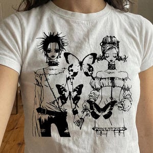 May include: White t-shirt featuring a black graphic of two figures holding butterflies. The figures have detailed hairstyles and clothing. The design is a stylized illustration with a gothic aesthetic.