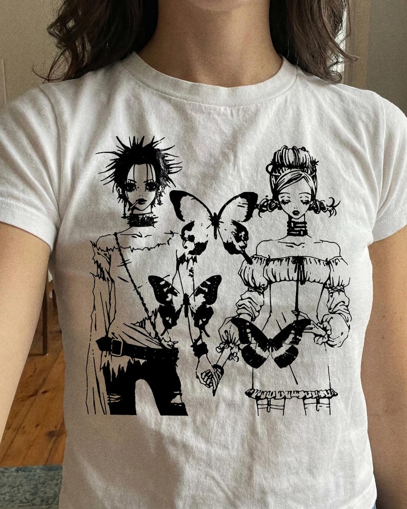May include: White t-shirt featuring a black graphic of two figures holding butterflies. The figures have detailed hairstyles and clothing. The design is a stylized illustration with a gothic aesthetic.