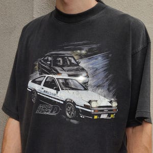Peut inclure: T-shirt gris foncé avec un motif de deux voitures de course. La voiture avant est blanche avec des détails noirs et du texte japonais. Le t-shirt a un aspect vintage et usé.