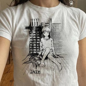 May include: White t-shirt featuring a black and white anime-style illustration of a seated figure with long hair, surrounded by wires and technology. The word "lain" is printed below the illustration.