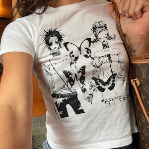 May include: White t-shirt featuring a black and white graphic of two anime-style figures and butterflies. The figures are dressed in detailed outfits. The shirt is paired with blue jeans.