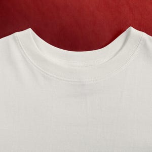 May include: Close-up of a white crew neck t-shirt. The neckline is ribbed and the fabric appears to be a soft cotton. The background is a solid red color.