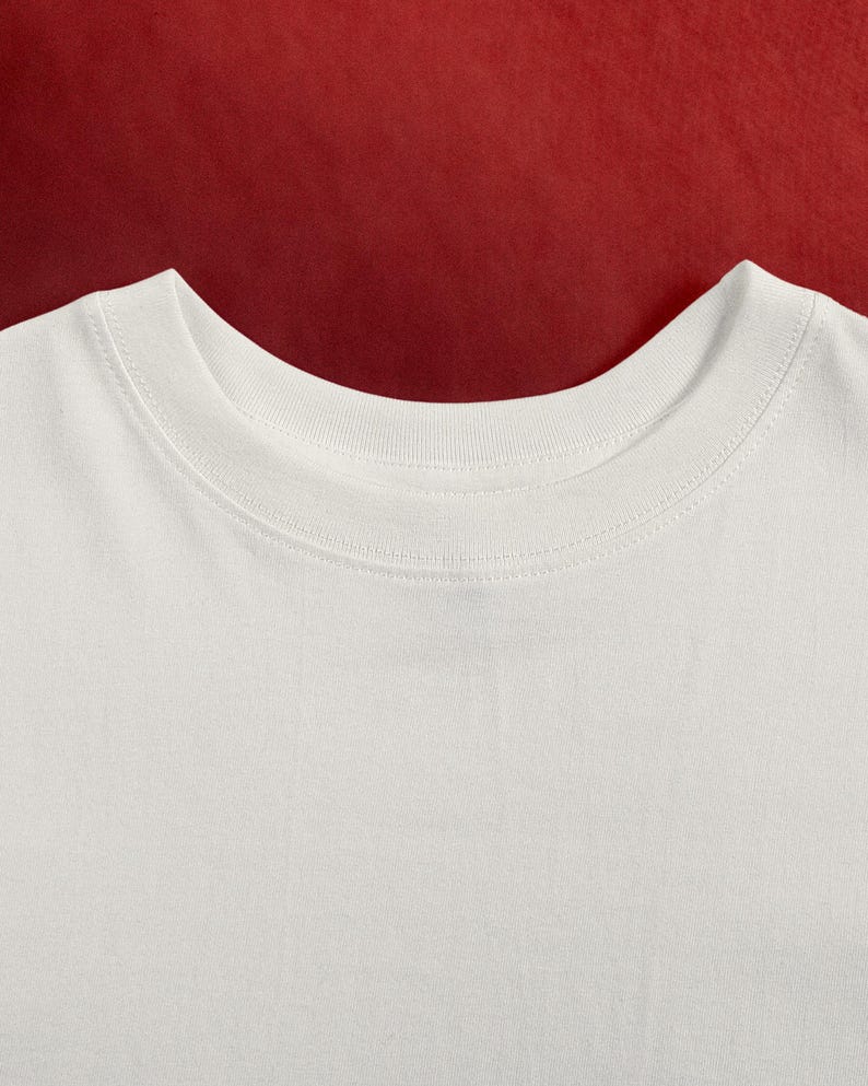 May include: Close-up of a white crew neck t-shirt. The neckline is ribbed and the fabric appears to be a soft cotton. The background is a solid red color.