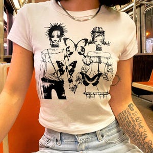 May include: White short-sleeve t-shirt featuring a black graphic of two figures with butterflies. The illustration has a manga style. The shirt is paired with light blue jeans.