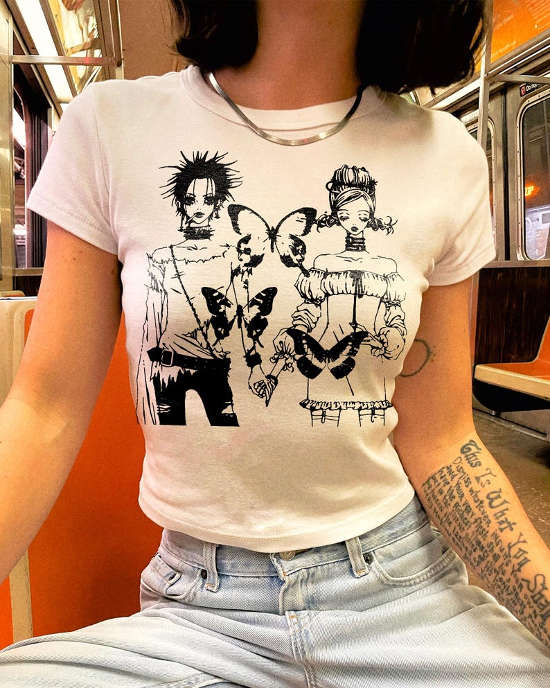May include: White short-sleeve t-shirt featuring a black graphic of two figures with butterflies. The illustration has a manga style. The shirt is paired with light blue jeans.