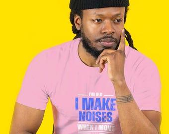 May include: A pink t-shirt with the text "I'M OLD I MAKE NOISES WHEN I MOVE" in blue and white. The person in the image is wearing a black beanie and has dreadlocks. The background is a bright yellow.