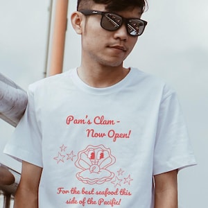 May include: White t-shirt featuring red text and graphics. The shirt displays "Pam's Clam - Now Open!" with a cartoon clam and the slogan "For the best seafood this side of the Pacific!" The address is also printed on the shirt.