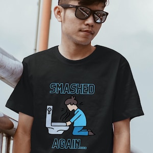 Könnte beinhalten: Schwarzes T-Shirt mit dem blauen Text "SMASHED AGAIN...". Die Grafik zeigt eine Person, die neben einer Toilette kniet. Die Person trägt blaue Kleidung und scheint sich zu übergeben. Die Toilette ist weiß.