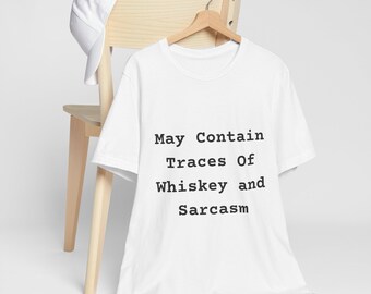 Könnte beinhalten: Weißes T-Shirt mit dem Text "May Contain Traces Of Whiskey and Sarcasm" in Schwarz. Das Shirt hängt an einem Holzkleiderbügel, mit einer weißen Baseballkappe auf einem Holzstuhl.