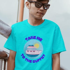Könnte beinhalten: Ein türkisfarbenes T-Shirt mit dem Schriftzug "TAKE ME TO THE BUFFET" in lila Schrift. Das Shirt zeigt eine Cartoon-Kreuzfahrt-Schiff- und Sonnengrafik in einem blauen Kreis. Die Person auf dem Bild trägt eine Sonnenbrille.