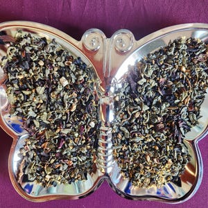 May include: A silver butterfly-shaped dish filled with loose leaf tea. The tea leaves are a mix of green, brown, and purple, with visible herbs and spices. The dish sits on a deep purple surface.