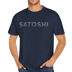 Può includere: T-shirt blu navy con la scritta "SATOSHI" stampata sul petto in uno stile digitale pixelato. La maglietta è a tinta unita e ha maniche corte e girocollo. Il modello indossa jeans blu.