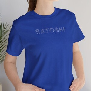 May include: Royal blue t-shirt with the word "SATOSHI" printed across the chest in a distressed, white font. The shirt is a crew neck style and is made of a soft material. The model is wearing blue jeans.