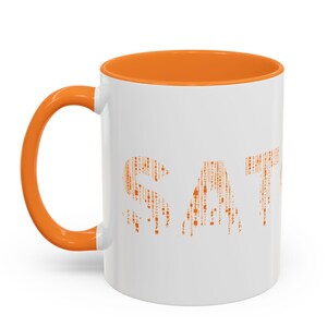 Può includere: Tazza in ceramica bianca con interno e manico arancione. La parola "SAT" è stampata in arancione sulla tazza, utilizzando un motivo di linee verticali e piccole forme. La tazza è progettata per bevande calde.