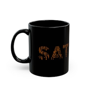 Può includere: Tazza da caffè in ceramica nera con manico curvo. La parola "SAT" è stampata sul lato in arancione, con carattere pixelato. La tazza è progettata per bevande calde.