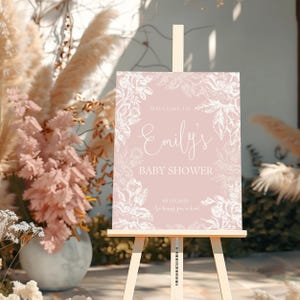May include: A pink baby shower welcome sign with white floral designs and the text "Welcome to Emily's Baby Shower 05.01.2023 So happy you're here!"