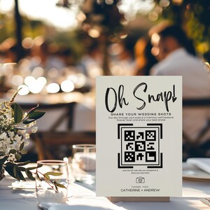 May include: A white sign with black text that reads "Oh Snap! Share Your Wedding Shots Our day through your eyes is a memory we'll treasure forever. Scan and share your best shots." A black and white QR code is below the text. Below the QR code is a camera icon and the text "Scan the QR code with your smartphone & share your best shots!" Below that is the text "Thank You Catherine + Andrew".
