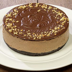 Puede incluir: Un pastel de chocolate redondo sobre un plato blanco, sobre un fondo de madera. El pastel tiene una cobertura de ganache de chocolate negro con un diseño en espiral y está espolvoreado con nueces picadas. El pastel se asienta sobre una corteza oscura y desmenuzable.