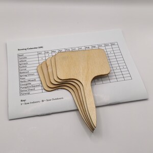 K&ouml;nnte beinhalten: Ein Stapel unfertiger Pflanzenmarkierungen aus Holz mit T-f&ouml;rmigem Griff, auf einem Aussaatkalender. Die Markierungen sind hellbraun und bereit zur individuellen Gestaltung. Der Kalender listet Gem&uuml;se und Kr&auml;uter auf.