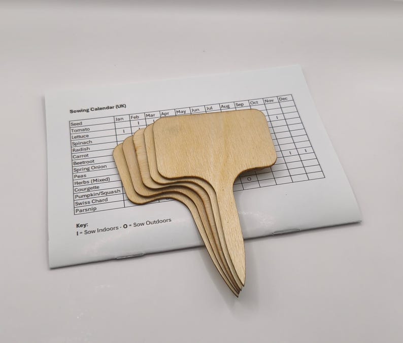 K&ouml;nnte beinhalten: Ein Stapel unfertiger Pflanzenmarkierungen aus Holz mit T-f&ouml;rmigem Griff, auf einem Aussaatkalender. Die Markierungen sind hellbraun und bereit zur individuellen Gestaltung. Der Kalender listet Gem&uuml;se und Kr&auml;uter auf.
