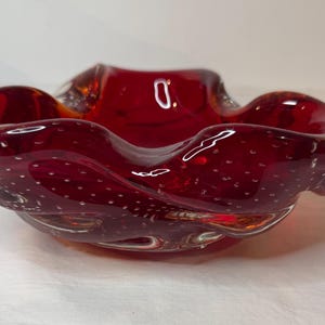 Peut inclure: Un bol en verre rouge rubis avec une forme ondulée et organique. Le bol présente une surface texturée avec de petites bulles d'air blanches. Le bord est légèrement évasé et le verre a une finition brillante. Ce bol décoratif est adapté à la décoration intérieure.