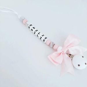May include: A pink and white silicone baby teether with the name "EMILIE" spelled out in black letters. The teether has a pink bow and a white clip.