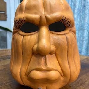 May include: A molded orange pumpkin mask featuring a stern, human-like facial expression with deep vertical ridges, a prominent nose, and dark, hollow eye sockets framed by long, black eyelashes. A small green stem sits atop the pumpkin head.