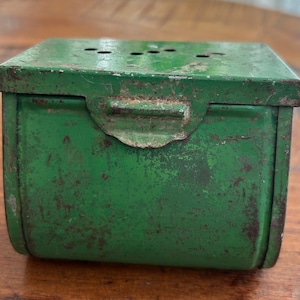 May include: A rectangular green metal box with a hinged lid featuring a series of small ventilation holes on the top surface. The exterior finish shows significant signs of wear, including patches of rust and chipped paint across the metal surface.
