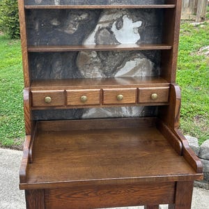May include: A tall wooden desk featuring a flat work surface, four small drawers with round brass-colored knobs, and three open shelves above. The back panel of the shelving unit displays a dark, marbled, and swirling gray and white pattern.