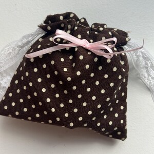 Könnte beinhalten: Eine braune Stofftasche mit Kordelzug, weißen Punkten und einem rosa Band, das oben gebunden ist. Die Tasche hat einen weißen Spitzenbesatz um die Öffnung.
