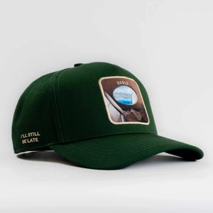 May include: A dark green baseball cap featuring a square patch on the front with an image of an airplane window view and the word OVALS. The side of the cap has the text I'LL STILL BE LATE embroidered in a light tan color.