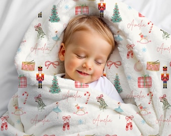 Könnte beinhalten: Eine weiße Babydecke mit einem Weihnachtsmuster, das Nussknacker, Geschenkpakete, Weihnachtsbäume und Ballettschuhe zeigt. Der Name "Amelia" ist in rosa Schrift über die Decke gedruckt.