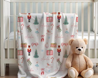 Könnte beinhalten: Eine weiße Babydecke mit einem sich wiederholenden Muster aus weihnachtlichen und Ballettthemen. Das Design umfasst Nussknacker, Weihnachtsbäume, Geschenkboxen, Balletttänzerinnen und den Namen "Amelia". Ein Teddybär sitzt neben der Decke.