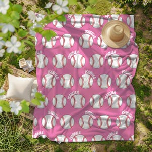May include: A pink blanket featuring a repeating pattern of baseballs and the name "Isabella" in white. A straw hat is placed on the blanket. The blanket is on grass.