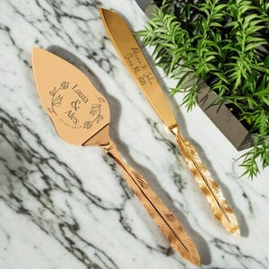以下が含まれることがあります： 羽根の形をしたハンドルが付いた、金色のウェディングケーキナイフとサーバーのセット。ナイフには「Alice & John June 11, 2023」と刻印されています。サーバーには「Laura & Alex 01.05.2023」と花柄が刻印されています。