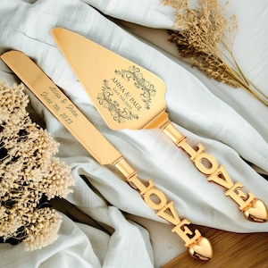 以下が含まれることがあります： 「LOVE」と刻まれた金色のウェディングケーキサーバーセット。サーバーの刃には、「Anna & Paul Save the Date」と「Alicia & John June 7th, 2023」の文字が刻まれています。