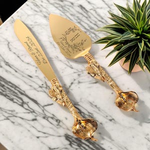 以下が含まれることがあります： 花柄のデザインとサーバーに刻まれた「H」と「R」のイニシャルが施された、金色のウェディングケーキナイフとサーバーのセット。ナイフのハンドルは、カサブランカの形をしています。サーバーのハンドルは、カサブランカの形をしています。ナイフには「Hena & Rumel Dec 22nd, 2024」という文字が刻まれています。