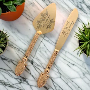 以下が含まれることがあります： 金色のウェディングケーキナイフとサーバーのセット。サーバーには花柄の彫刻と「K & L」と「June 22nd, 2024」の名前が、ナイフには「Allison & John June 22nd, 2023」の名前が刻まれています。