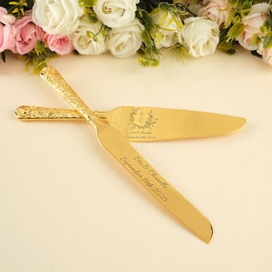 以下が含まれることがあります： 金色のケーキナイフとサーバーのセット。ハンドルはテクスチャード加工のデザインで、ブレードには名前と日付が刻印されています。背景には花の配置が施されています。