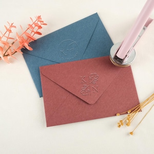 以下が含まれることがあります： エンボス加工が施された、ダスティーローズとブルーの2つの封筒。ダスティーローズの封筒には、花のアクセントが付いたイニシャル「K」と「N」があります。青い封筒にはイニシャルと日付があります。ピンクのホッチキスが上にあります。